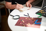 A jeweler takes notes while evaluating gold jewelry on a workbench with tools, a loupe, and several pieces laid out.