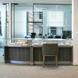 A modern jewelry display counter with glass cases and a gray upholstered chair, set in a bright office interior with large glass walls and windows.