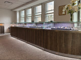 A long curved jewelry display case with wood-grain fronts and glass tops, set in a showroom with large windows, carpeted floor, and a vase of white flowers on the wall.